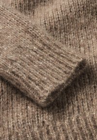 Brauner Strickpullover aus einem Material mit Rippstruktur und aufgerolltem Ärmelabschluss, der weiche, flauschige Fasern und ein warmes Erscheinungsbild zeigt.