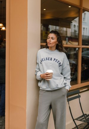 Szara bluza z napisem "Chelsea Peers", w połączeniu z jasnoszarymi spodniami. Trzyma kubek z kawą, stoi przy beżowej ścianie.