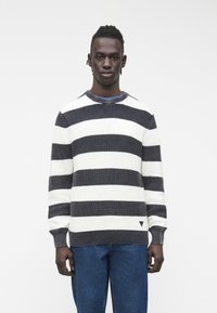 Moški, oblečen v črno-bel pleten pulover s širokimi črtami in modre kavbojke, stoji na preprostem svetlem ozadju.