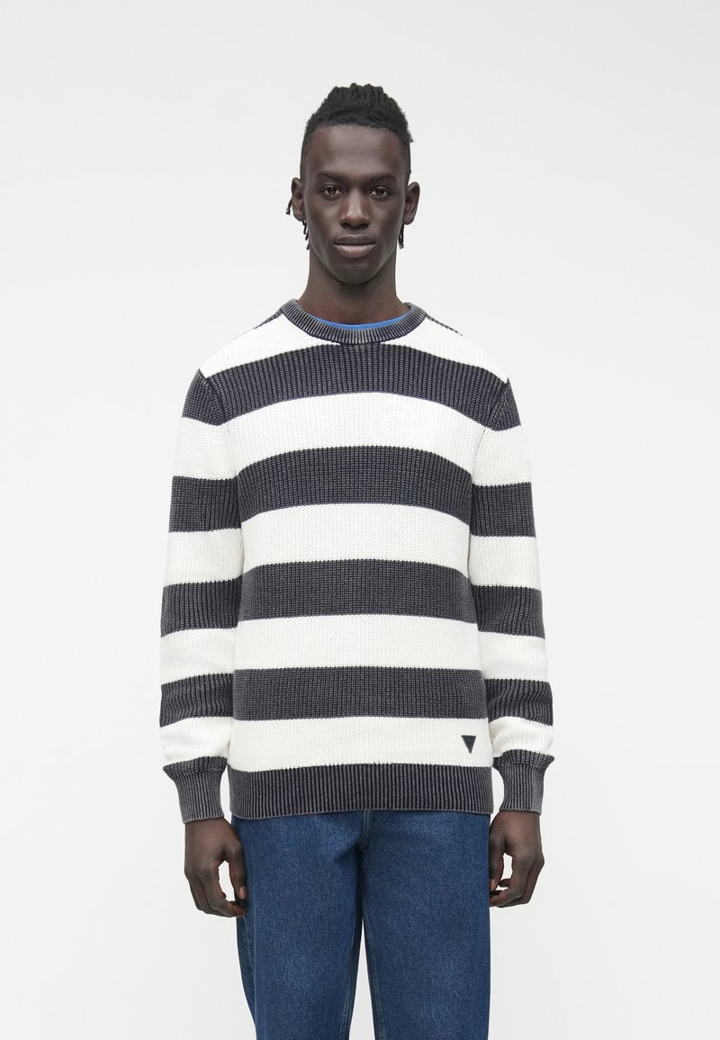 Moški, oblečen v črno-bel pleten pulover s širokimi črtami in modre kavbojke, stoji na preprostem svetlem ozadju.