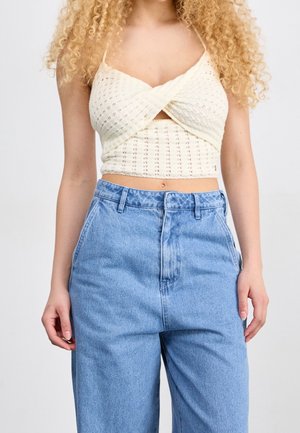Kobieta nosząca kremowy dzianinowy crop top z krzyżującym się przodem i jasnoniebieskie, wysokie na talii dżinsy na jednolitym białym tle.