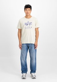 Krátký rukáv béžové tričko s modrým potiskem loga, kombinované s světle modrými džíny a šedými teniskami. Jednoduchý design, uvolněný střih.