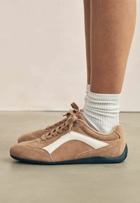 Baskets en daim beige avec des accents blancs, design à lacets, bout arrondi et semelle en caoutchouc noir, associées à des chaussettes blanches à côtes.