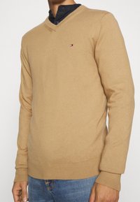 Pull en maille couleur chameau avec col en V, manches longues, poignets et ourlet côtelés. Présente un petit logo sur la poitrine. Texture douce en maille.