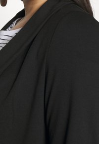 Blazer noir en tissu doux et lisse. Présente un design drapé, une ouverture sur le devant et un col replié, avec un haut rayé visible en dessous.