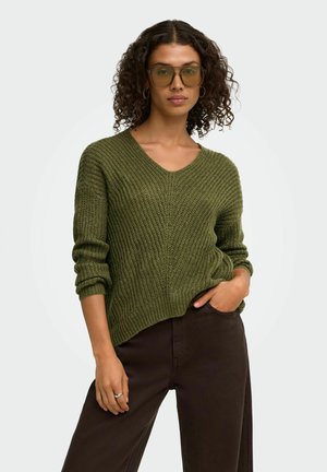 Femme aux cheveux bouclés portant un pull en maille vert olive, un pantalon marron, des lunettes teintées jaunes et des boucles d'oreilles créoles argentées, posant avec une main dans la poche.