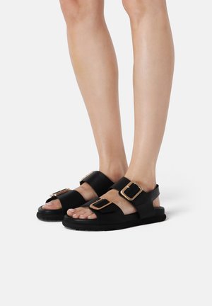 Zwarte sandalen gemaakt van glad materiaal met twee brede banden en grote, vierkante gouden gespen. Platte zool met een gestructureerde zwarte rand.