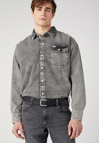 Grijze denim overhemd met een knoopsluiting, borstzak en contrasterende stiksels, gecombineerd met donkere grijze jeans en een zwarte riem.