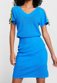 Femme portant une robe tricotée bleu vif à manches courtes avec des détails de ruban rayé jaune sur les épaules, posant devant un fond uni.