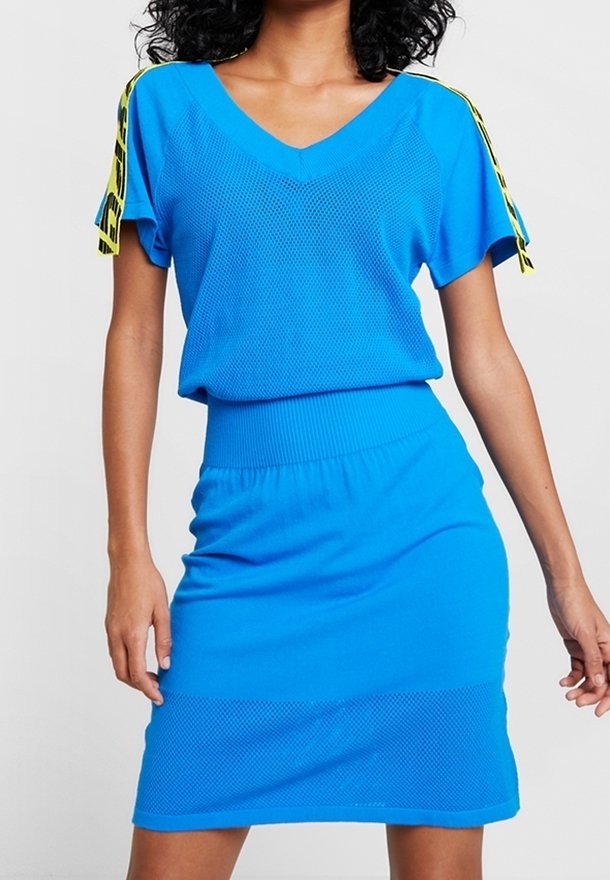 Femme portant une robe tricotée bleu vif à manches courtes avec des détails de ruban rayé jaune sur les épaules, posant devant un fond uni.