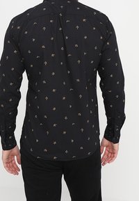 Zwarte shirt met kleine gouden geometrische patronen, met knoopmanchetten en een enkele rug yoke ontwerp. Zachte stof met een gladde textuur.