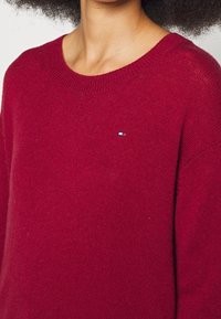 Femme portant un pull en maille rouge profond avec un petit logo de drapeau rectangulaire sur la poitrine, montrant les épaules et le cou.