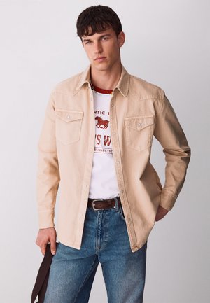 Giovane uomo che indossa una camicia beige con bottoni sopra una t-shirt bianca con grafica, jeans blu e cintura marrone, che tiene una cinghia scura nella mano sinistra.