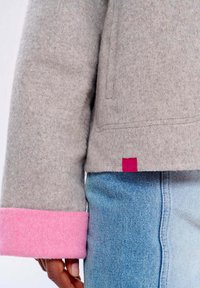 Šedá vlněná bunda s růžovými manžetami a odpovídající růžovou nášivkou. Má jednoduchý design a měkkou strukturu. Kombinováno s modrými džínami.