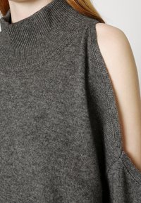 Šedý pletený svetr s vysokým límcem a otevřeným designem ramen. Měkká textura s žebrovanými detaily kolem límce a otvorů na pažích.