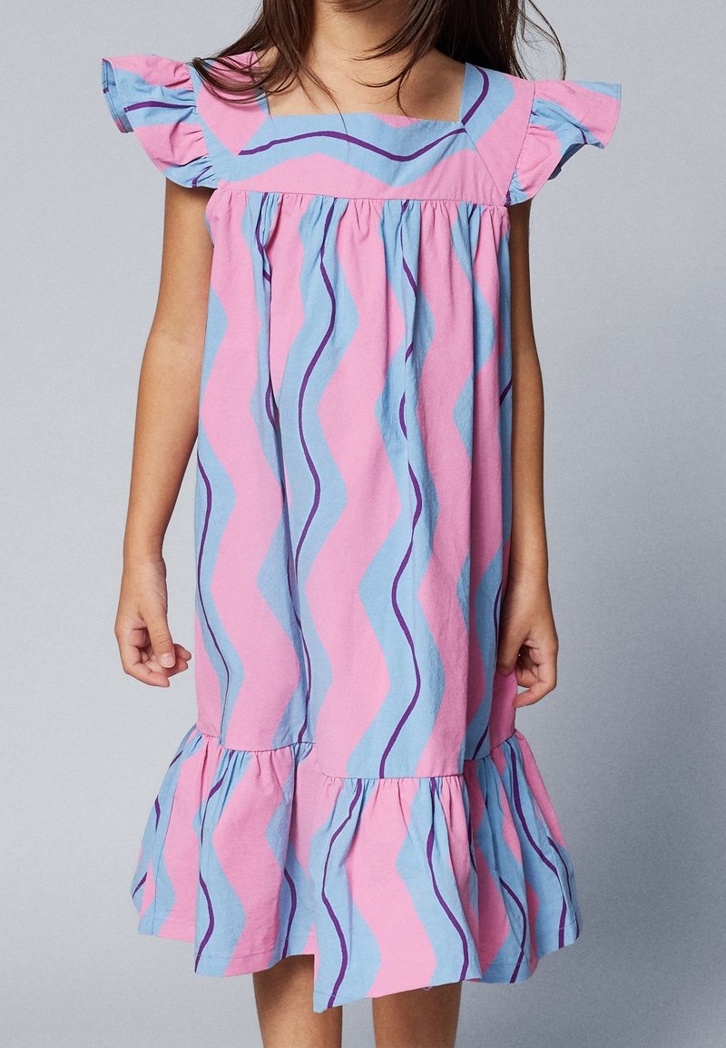 Enfant portant une robe rose sans manches avec des rayures ondulées bleues et violettes, des épaules à volants et un ourlet à volants, debout devant un fond uni.