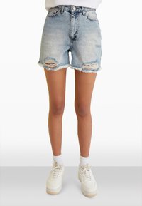 Pantalones cortos de mezclilla azul claro con dobladillo deshilachado, detalles rasgados y diseño ajustado. Combinados con zapatillas blancas y calcetines blancos.