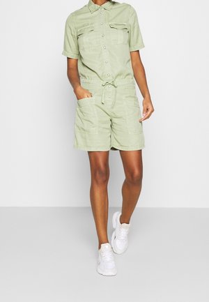 Lys grøn kortærmet jumpsuit med knaplukning, to brystlommer og bæltebinding i taljen; båret med hvide sneakers.