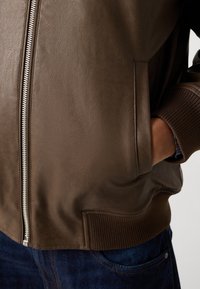 Giacca bomber in pelle marrone con finitura testurizzata, chiusura lampo argentata, polsini e fondo a coste, e tasche laterali. Jeans in denim visibili nella parte inferiore.