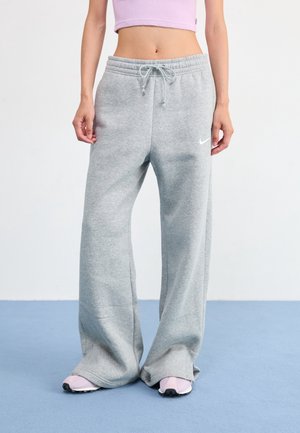 Person wearing loose-fitting gray sweatpants with drawstring waist and light pink sneakers standing on blue floor against white wall.