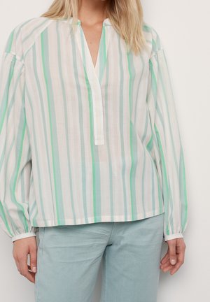 Frau trägt eine lockere, langärmelige Bluse mit vertikalen grünen und weißen Streifen, kombiniert mit hellblauen Hosen.