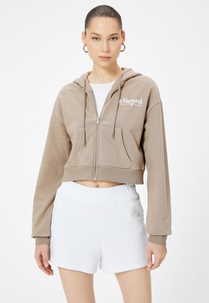 Felpa beige cropped con zip frontale, tasca a marsupio e stampa bianca di New York. Abbinata a pantaloni corti bianchi con vita elastica.