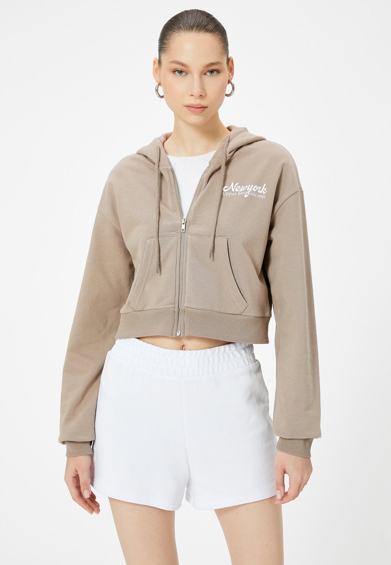 Felpa beige cropped con zip frontale, tasca a marsupio e stampa bianca di New York. Abbinata a pantaloni corti bianchi con vita elastica.