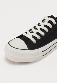 Sneaker nera in tela con suola in gomma bianca, punta rotonda e lacci bianchi a contrasto. Presenta una parte superiore testurizzata e strisce laterali.