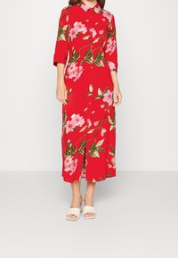 Abito midi floreale rosso con chiusura a bottoni, maniche a 3/4 e spacco frontale. Presenta fiori rosa e foglie verdi su un tessuto liscio. Indossato con sandali.