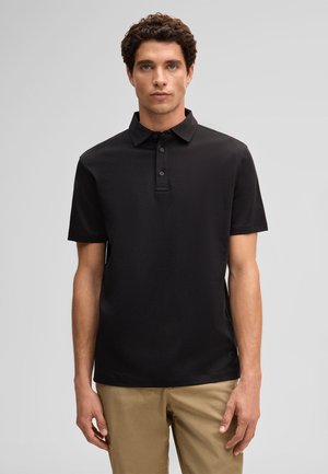 Junger Mann mit lockigem dunklem Haar, der ein schwarzes Poloshirt mit kurzen Ärmeln und beige Hose trägt, vor einem einfarbigen hellgrauen Hintergrund stehend.