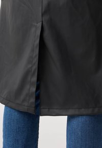 Czarny płaszcz przeciwdeszczowy o prostym kroju, z bocznymi rozcięciami i gładką, błyszczącą fakturą, zestawiony z niebieskimi jeansami.