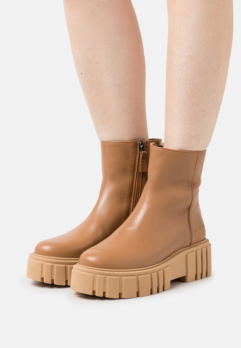 Bottes cheville en cuir fauve avec semelles plateformes épaisses et texturées, portées sur des jambes nues sur fond blanc.