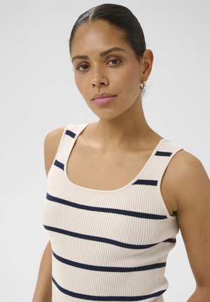 Kvinde med mørkt hår i en lav knold iført en cremet ribbet tanktop med horisontale marineblå striber, kigger let mod kameraet.