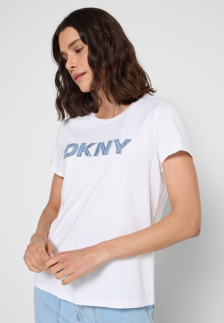 Camiseta blanca de manga corta con cuello redondo, que presenta letras "DKNY" en textura de denim azul en la parte delantera. Tejido suave y liso.