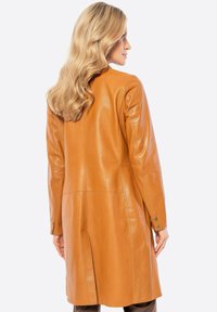 Tan leren jas met een rechte snit, lange mouwen en een gecentraliseerde naad. Beschikt over knoopdetails aan de manchetten en een achterventilatie voor bewegingsvrijheid.