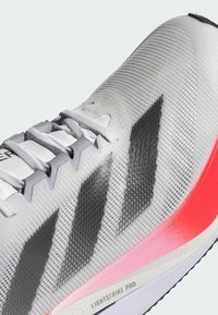Zapatilla deportiva blanca con una parte superior sintética texturizada, que presenta rayas negras y una suela rosa brillante marcada "LIGHTSTRIKE PRO."
