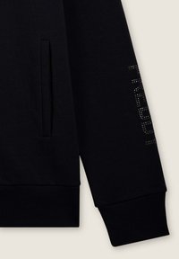 Sudadera negra con una tela suave y texturizada; presenta un bolsillo lateral y un acento de strass a lo largo de la manga. Los puños son acanalados.