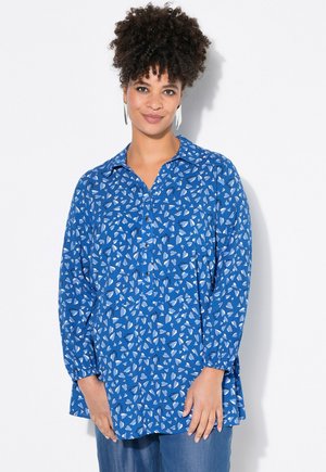 Blauwe blouse met lange mouwen in een lichte stof, versierd met een hartvormig patroon in wit en marineblauw. Met knopen aan de voorkant en een kraag.