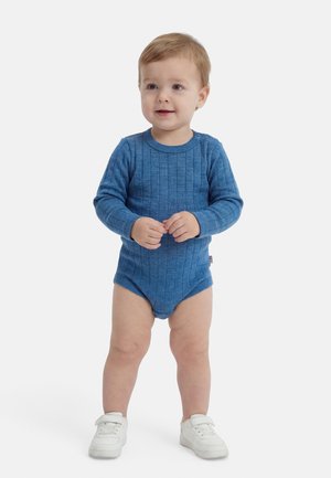 Tout-petit debout portant un body côtelé à manches longues bleu et des baskets blanches sur un fond blanc.