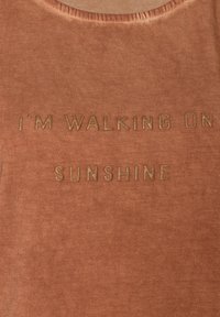 Rostfarbenes Gewebe mit weicher Textur, versehen mit goldener Metallicschrift "I'M WALKING ON SUNSHINE", die zentral auf der Vorderseite platziert ist. Runder Ausschnitt.