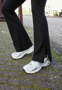 Crne flare hlače s detaljima na zatvaraču na rubu, u kombinaciji s bijelim i srebrnim New Balance tenisicama koje imaju mrežaste i gumene potplate.