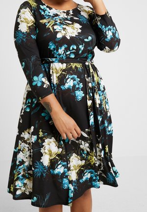 Femme portant une robe noire avec un motif floral bleu, blanc et vert, et une ceinture en tissu assortie nouée à la taille.