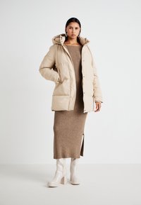 Doudoune beige avec capuche, dotée d'une fermeture éclair à l'avant et de boutons-pression. Portée sur une robe en tricot côtelé beige, associée à des bottes à talons légers.