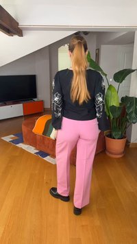 Kvinne med langt blondt hestehale, iført svart blomstret langermet topp og rosa bukser, stående i stuen med planter og møbler.