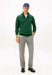 Groen gebreide trui met een kwartrits, grijze broek en zwarte schoenen. Model draagt een blauwe pet. Minimalistische, casual outfit getoond.