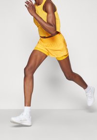 Gelbes sportliches Tanktop und Shorts aus einem leichten Material. Die Shorts haben einen elastischen Bund; das Modell trägt weiße Sneakers und Socken.