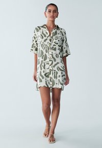 Abito camicia a maniche corte, con bottoni, di colore beige chiaro con motivi astratti verdi. Presenta una vestibilità comoda e arriva sopra il ginocchio.