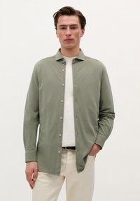 Cămașă lungă cu mânecă verde deschis, cu guler căptușit și butoane pe față, asortată cu un tricou alb și pantaloni bej. Textură netedă.