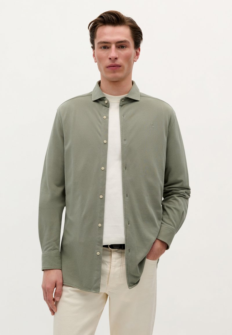 Cămașă lungă cu mânecă verde deschis, cu guler căptușit și butoane pe față, asortată cu un tricou alb și pantaloni bej. Textură netedă.