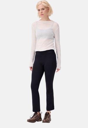 Blonde Frau trägt ein durchsichtiges weißes Langarmoberteil, schwarze hoch taillierte eng anliegende Hose und braune Schnürsneaker, steht vor weißem Hintergrund.